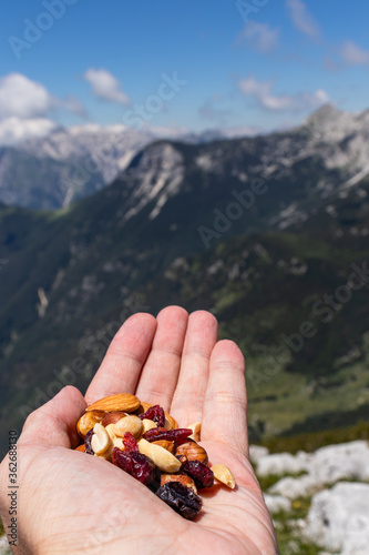 Hand holding a healthy snack on the top of the mountain after climbing. Consisting of different nuts and dried fruit, with beautiful background of mountain and forest in the summer. 
