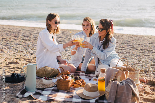Three young beautiful happy women female friends having cozy summer picnic with lemonade, fresh bread and fruits on a beach.