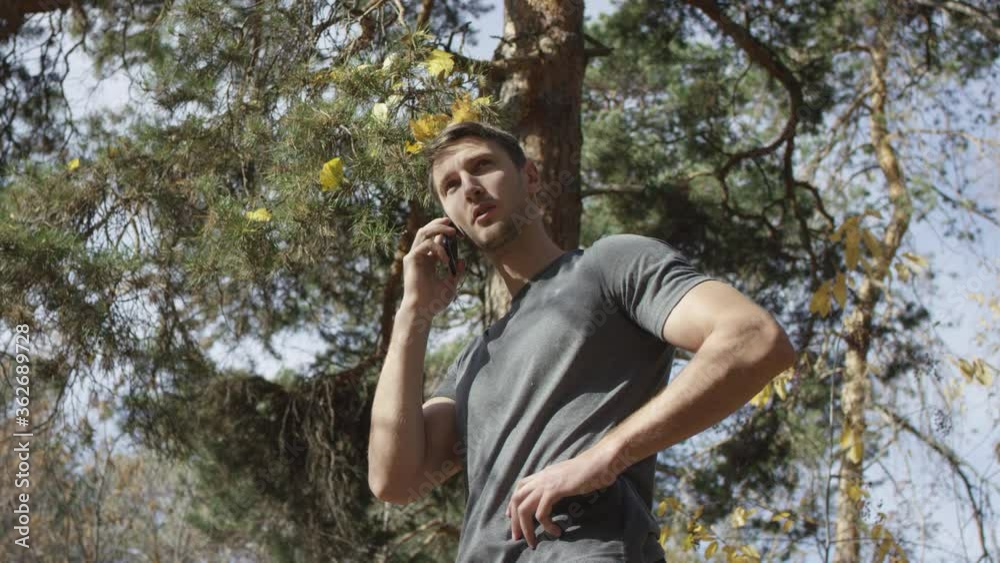 Good-looking thoughtful male in grey shirt has important phone call and listens patiently while standing in autumn sunlit forest. Low angle 4K 360 degree tracking arc shot. Good-looking thoughtful male in grey shirt has important phone call and listens patiently while standing in autumn sunlit forest. Low angle 4K 360 degree tracking arc shot.