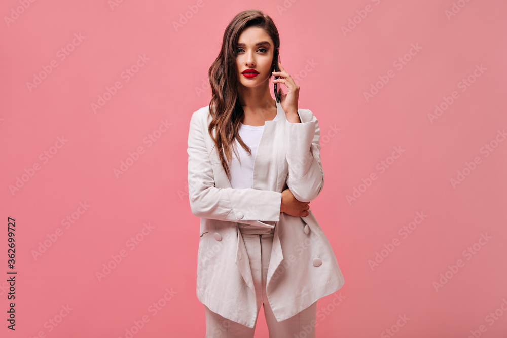 Curly woman in white suit talks on phone. Stylish lady with red lips in light fashionable outfit on pink isolated background..