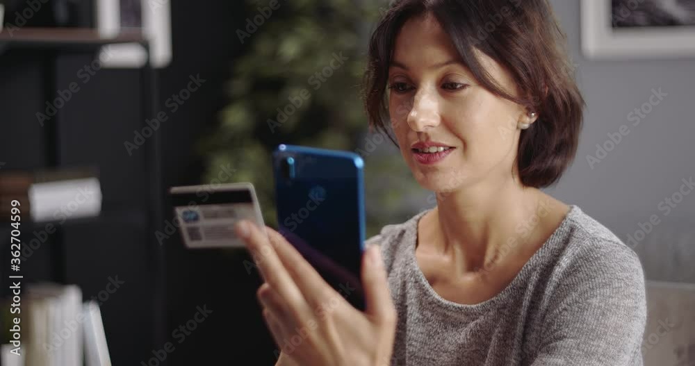Attractive woman with brown hair using smartphone and credit card for online payments. Happy housewife sitting at living room and doing shopping on modern gadget. Attractive woman with brown hair using smartphone and credit card for online payments. Happy housewife sitting at living room and doing shopping on modern gadget.