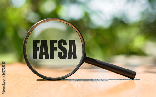 Free Application for Federal Student Aid - FAFSA written on magnifying glass on wooden table and green background. Concept image