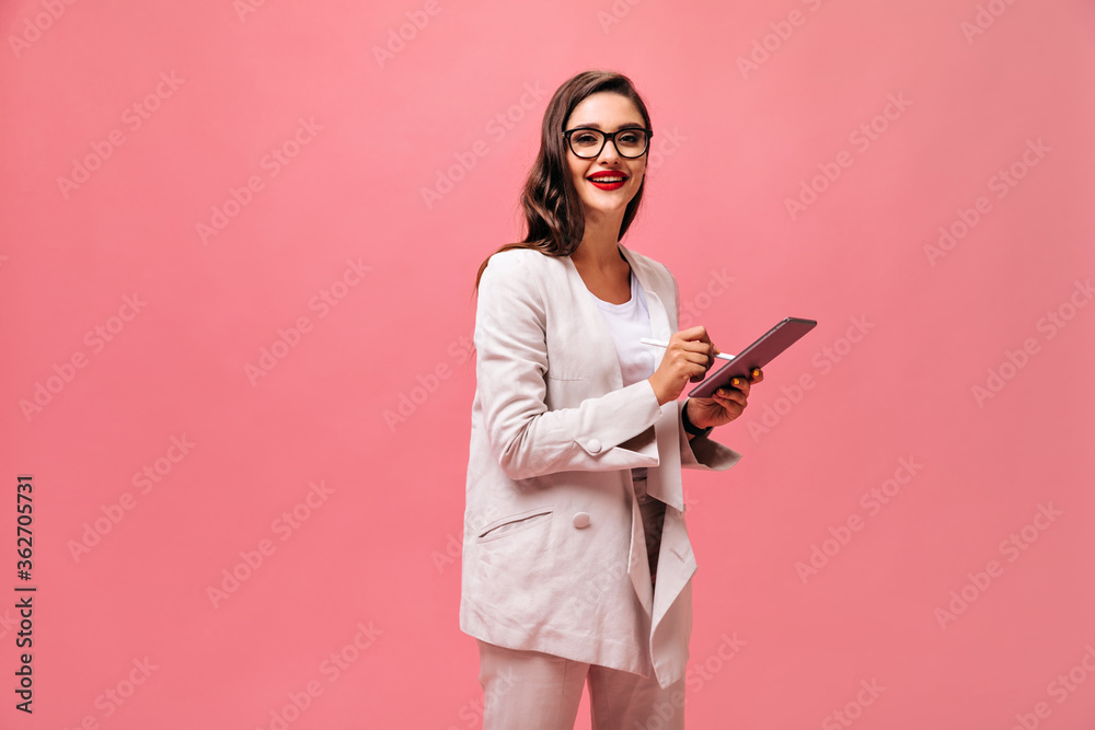 Pretty wonderful woman with red lips in fashionable jacket and eyeglasses poses with computer tablet on isolated pink background..