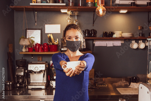 Caucasian woman offering coffee cup with both hands in a small business wearing mask durind covid-19 pandemic reopening after quarantine photo