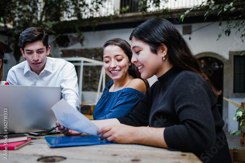 Canvas-taulu Young latin woman working with computer and her coworkers at the office or cowor