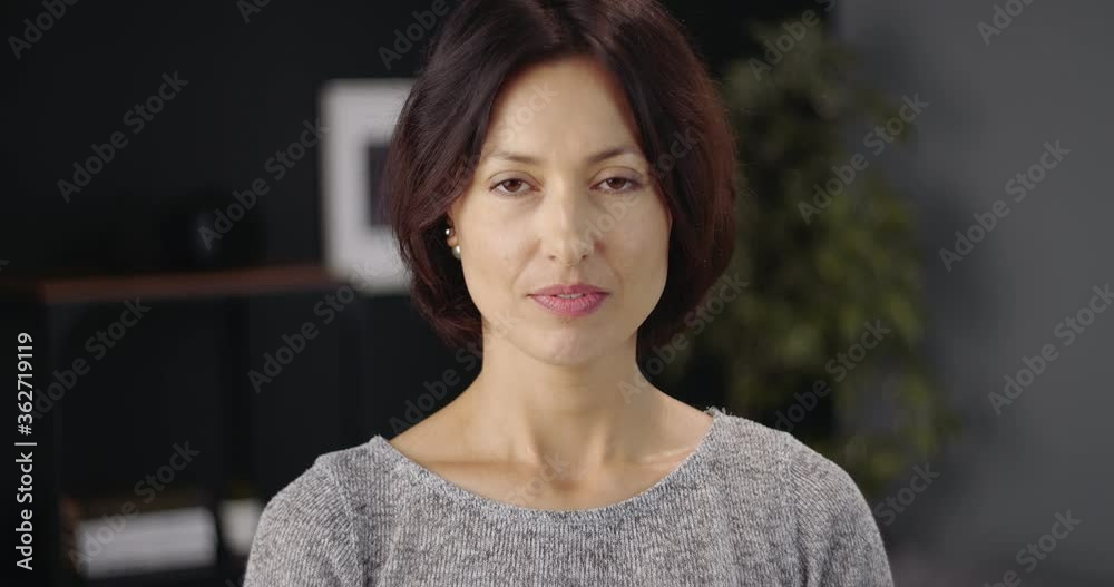 Portrait of attractive woman with dark hair standing in modern apartment and posing on camera. Mature lady in casual outfit smiling and looking ahead. Portrait of attractive woman with dark hair standing in modern apartment and posing on camera. Mature lady in casual outfit smiling and looking ahead.