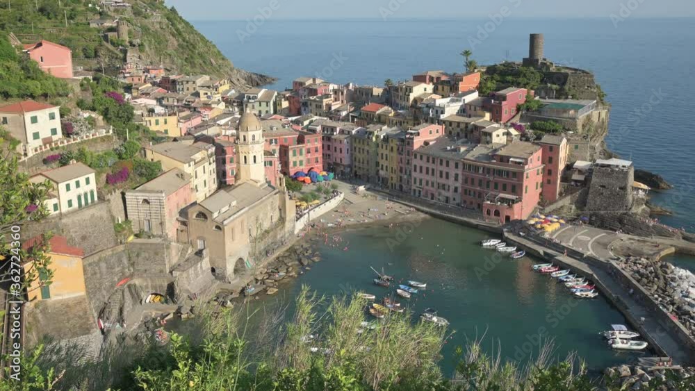 Vernazza,Liguria,Italy.June 2020. Beautiful view of the village: in the center the small port with its crystal clear sea,around the houses with colored facades.People walk in the square. Tilt movement Vernazza,Liguria,Italy.June 2020. Beautiful view of the village: in the center the small port with its crystal clear sea,around the houses with colored facades.People walk in the square. Tilt movement
