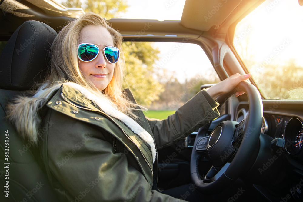 Naklejka premium Blonde female driver, driving in nature in convertible with aviator sunglasses and jacket, during sunset