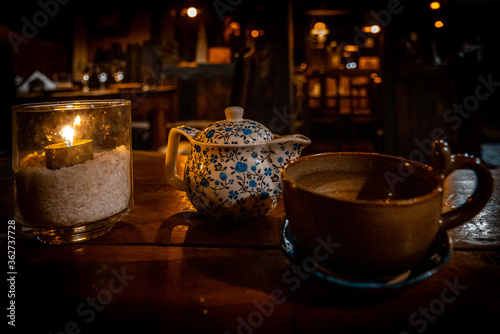 A teapot, a cup and a tealight inside a glass illuminating these, over a wooden table of a cozy Irish pub