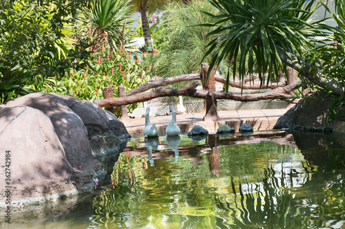 The bridge has 2 duck statues. There are wood around the garden.