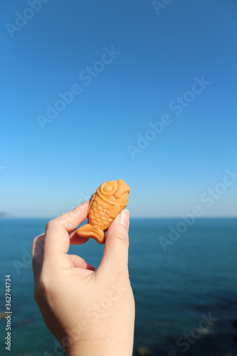 Hand holding a mini fish-shaped waffle with custard filling with the sea background in sunny day, South Korea © Crystaltmc