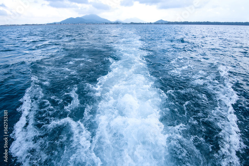 Trail and foamy waves on blue sea surface behind of moving boat with island background.
