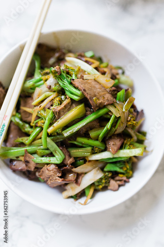 A delicious bowl with stir-fried beef and broccoli in a round white porcelain bowl with chopsticks resting on the bowl. Vertical photo.
