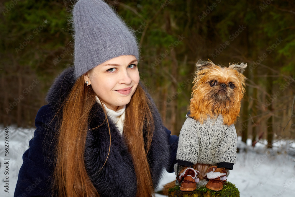 Girl with long hair in black jacket and grey hat posing with a small dog Brussels Griffon in clothes in the forest or park in winter day Girl with long hair in black jacket and grey hat posing with a small dog Brussels Griffon in clothes in the forest or park in winter day