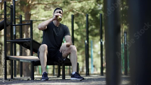 Adult caucasian strong sportsman is taking a rest on a bench and drinks water out of the shaker at the gym at the park