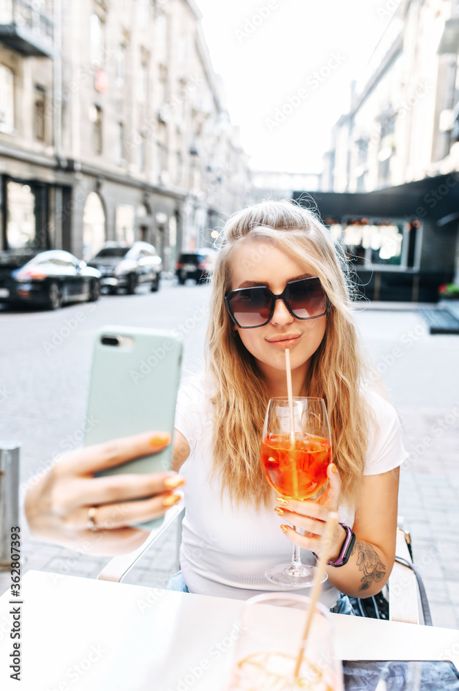 Fancy blonde girl sunglasses drinks a cocktail and takes a on the trendy smartphone while sitting outdoor in the cafe. Vertical shot Stock-foto | Adobe Stock