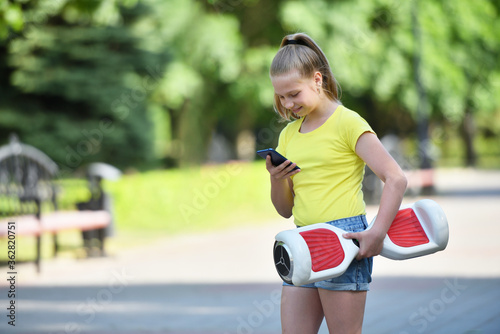 Smiling girl child used a smartphone while walking in a park and holding a hoverboard, authentic photo