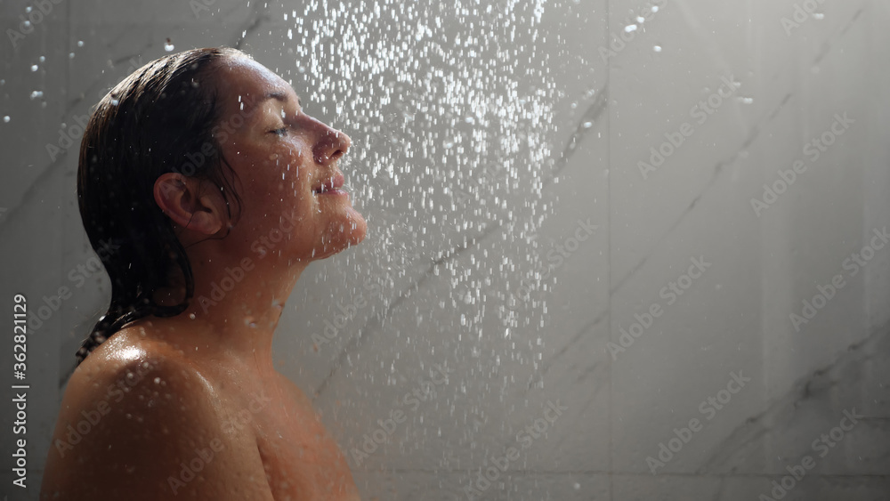 pretty brunette with bare shoulders and long wet hair stands under shower jets behind bathroom glass copy space