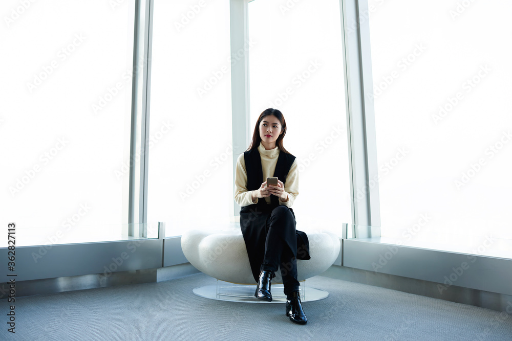 Fototapeta premium Young Asian female is thinking that to write in text message on cell phone,while is waiting for deparure of flight in airport hall. Businesswoman is sitting in modern interior of big skyscraper office