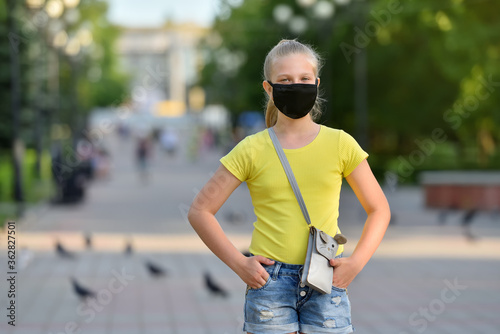 Girl child in a black protective mask against the backdrop of a summer park during the COVID-19 pandemic looks sad at the camera