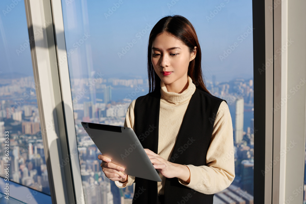 Japanese woman professional worker in real estate company is reading advertising in internet via digital tablet, while is standing in modern office interior near window with view of business district Japanese woman professional worker in real estate company is reading advertising in internet via digital tablet, while is standing in modern office interior near window with view of business district
