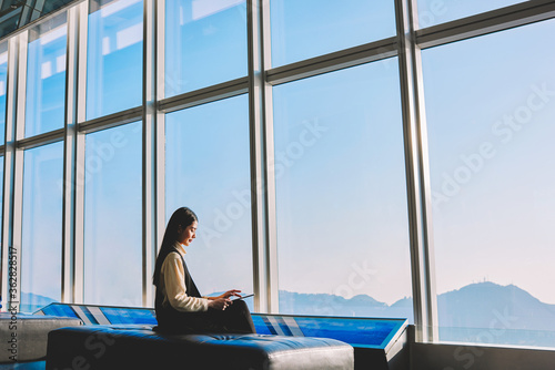 Young confident Asian female employer is using digital tablet for searching needed information during work break, while sitting in office interior against big window with cityscape view on background
