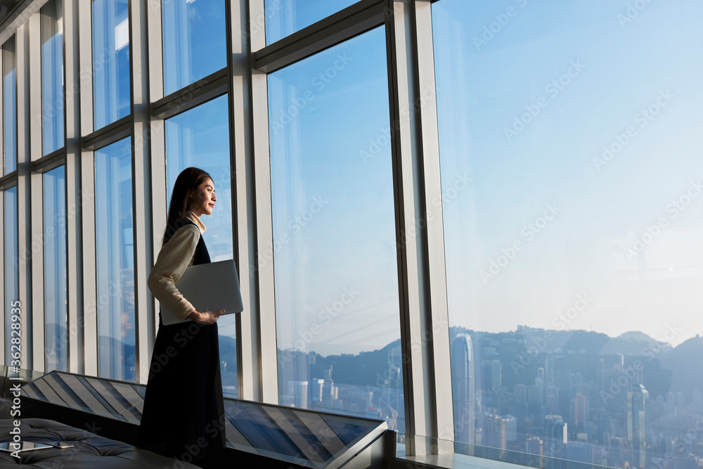 Chinese successful and satisfied businesswoman is standing with laptop computer in modern office interior near skyscraper window with copy space area for advertise text message or promotional content