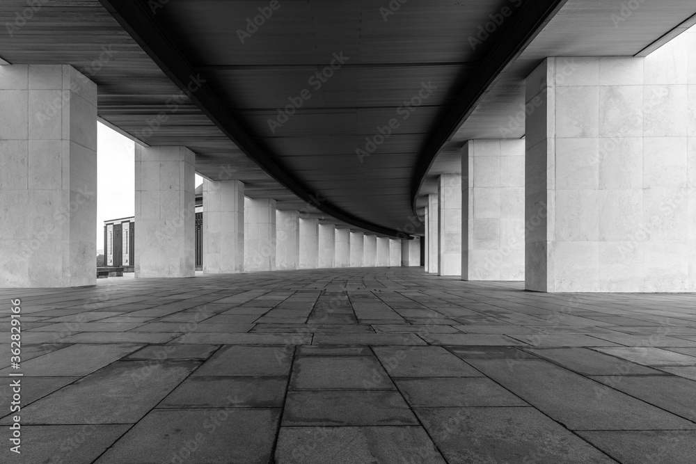 A smoothly curved gallery with rectangular vertical light columns of stone. Dark ceiling and floor. A distant perspective. Black and white image.