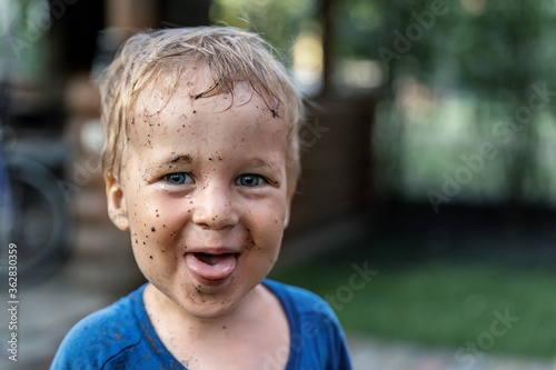 Cute adorable blond caucasian little happy toddler boy portrait with messy mus spots on face after playing watering garden at yard or countyside farm. Childhood happiness summertime country concept photo