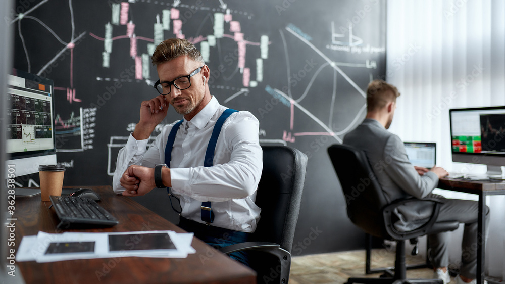 Earn right. Stylish businessman, trader looking at his wristwatch while working using wireless earphones. His colleague and blackboard full of charts and data analyses in the background. Earn right. Stylish businessman, trader looking at his wristwatch while working using wireless earphones. His colleague and blackboard full of charts and data analyses in the background.