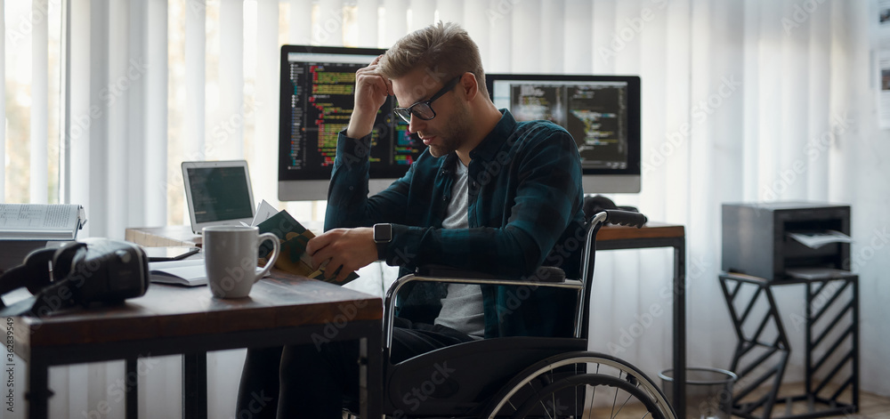 More knowledge. Portrait of concentrated male web developer in a wheelchair reading a book while sitting at his workplace in the office. Multiple computer screens with program code More knowledge. Portrait of concentrated male web developer in a wheelchair reading a book while sitting at his workplace in the office. Multiple computer screens with program code