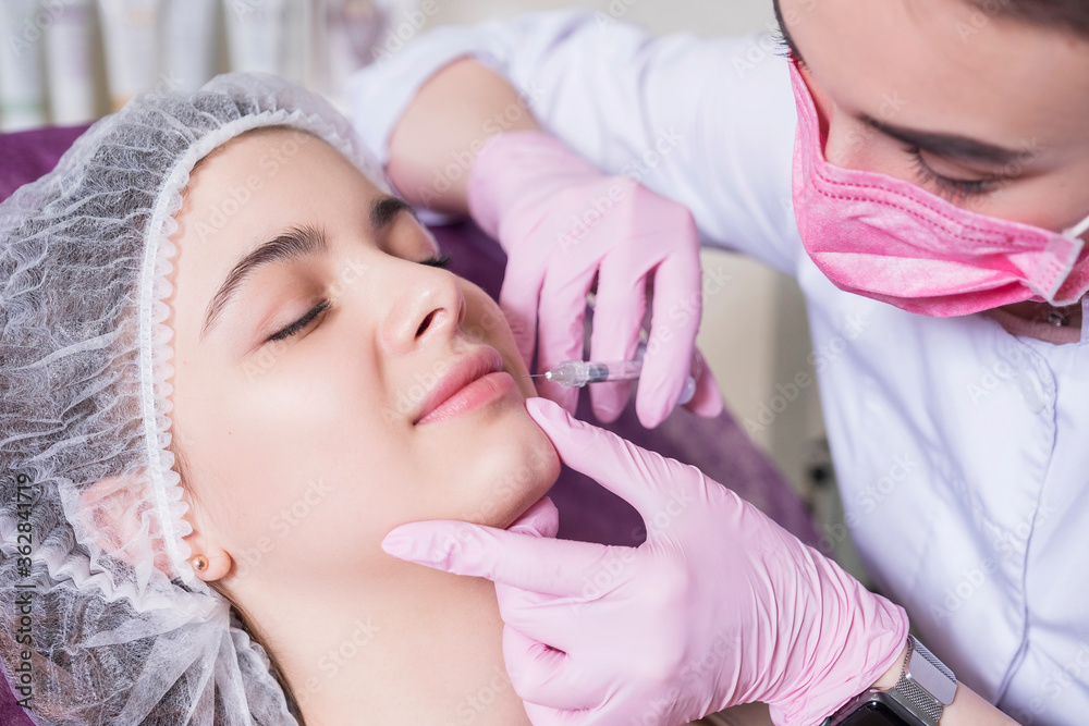 Cosmetologist doctor do injection procedure to a woman in her clinic. She wear glowes and mask and holds syringe with a needle Cosmetologist doctor do injection procedure to a woman in her clinic. She wear glowes and mask and holds syringe with a needle