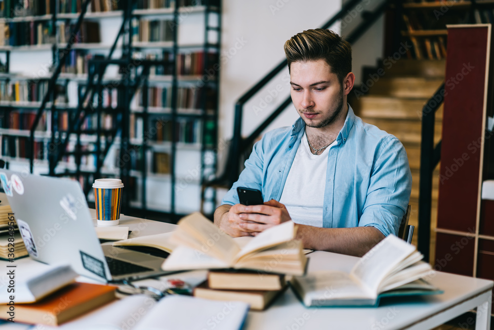 Thoughtful man browsing smartphone at table full of books and laptop Thoughtful man browsing smartphone at table full of books and laptop