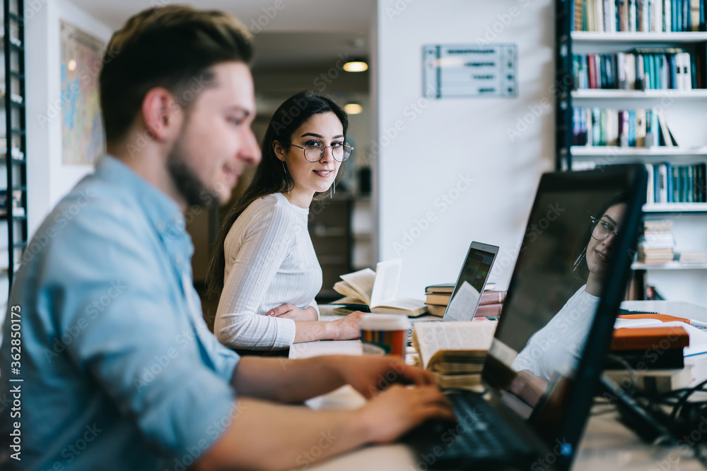 Portrait of skilled female employee looking at camera sitting next to colleague on blurred frontage, young male and female students using modern laptops for research and studying online course