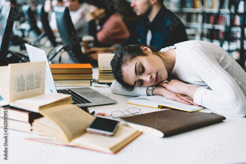 Tired female hipster in casual wear sleeping on table with books at library