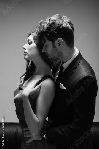 Monochrome image of handsome man in suit kissing and embracing beautiful woman in dress on grey