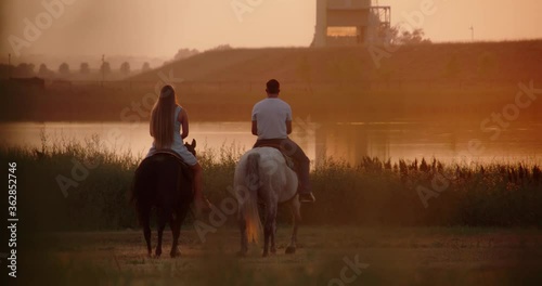 Caucasian couple ride horses during golden hour on farmland by a beautiful lake. Long lens handheld slow motion shot. 