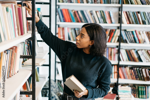 Young female african american librarian making order on bookshelves in college, dark skinned woman student picking literature for education checking information in books store, hipster girl get novels