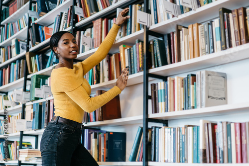 Portrait of attractive dark skinned young woman choosing book from bookshelf while smiling at camera.Charming african american student pulling literature textbook in library to preparing for exams