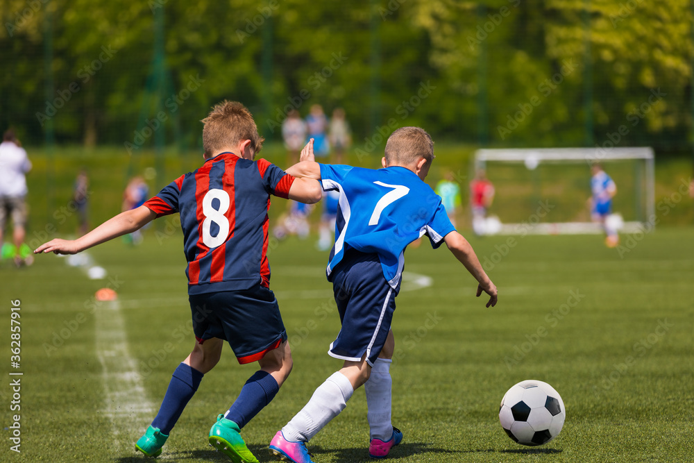 Two youth football players running in duel after ball. Kids kicking soccer match on the grass field. School children in sports jersey clothes with numbers on back in tough competition Two youth football players running in duel after ball. Kids kicking soccer match on the grass field. School children in sports jersey clothes with numbers on back in tough competition
