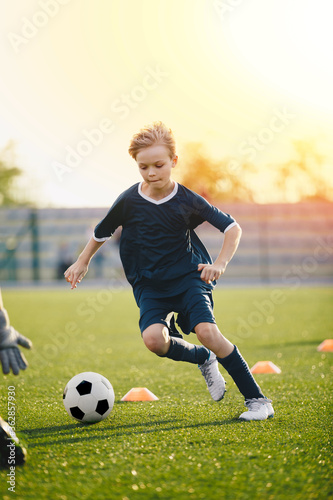 Young blonde boy kicking soccer ball on drill. Kids playing football training game on the school sports pitch. Soccer stadium and training equipment covered by summer sunset in the background