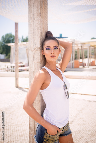 Free Download Sexy Caucasian Woman on the Beach With White Undershirt, Jeans Shorts, Sunglasses and Pony Tail, Leaning on a Wooden Pole, Looking Straight - GFXLoad