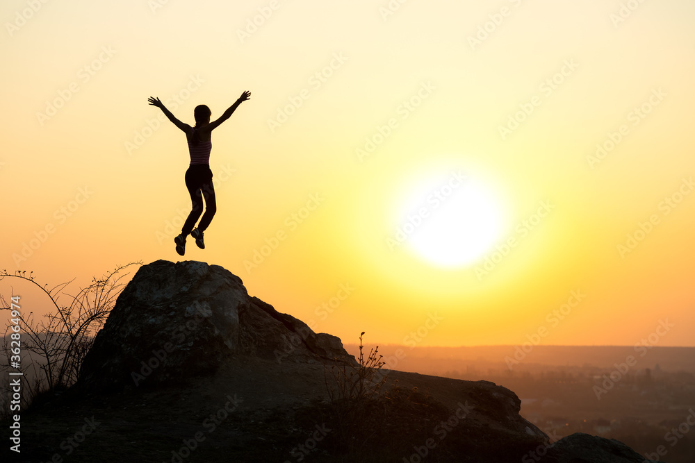 Silhouette of woman hiker jumping alone on empty rock at sunset in mountains. Female tourist raising her hands up standing on cliff in evening nature. Tourism, traveling and healthy lifestyle concept.