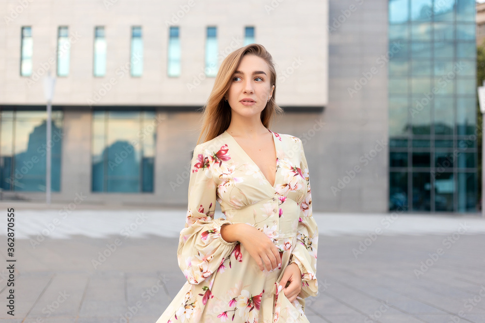 Portrait of a beautiful young girl on a background of a blue business center dressed in a summer beige dress