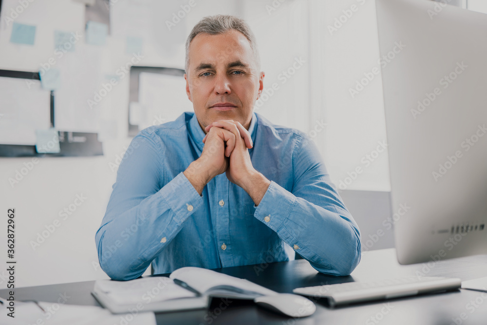 handsome gray-haired man sits in his office holding both hands near face while working on business project looks satisfied, work routine concept