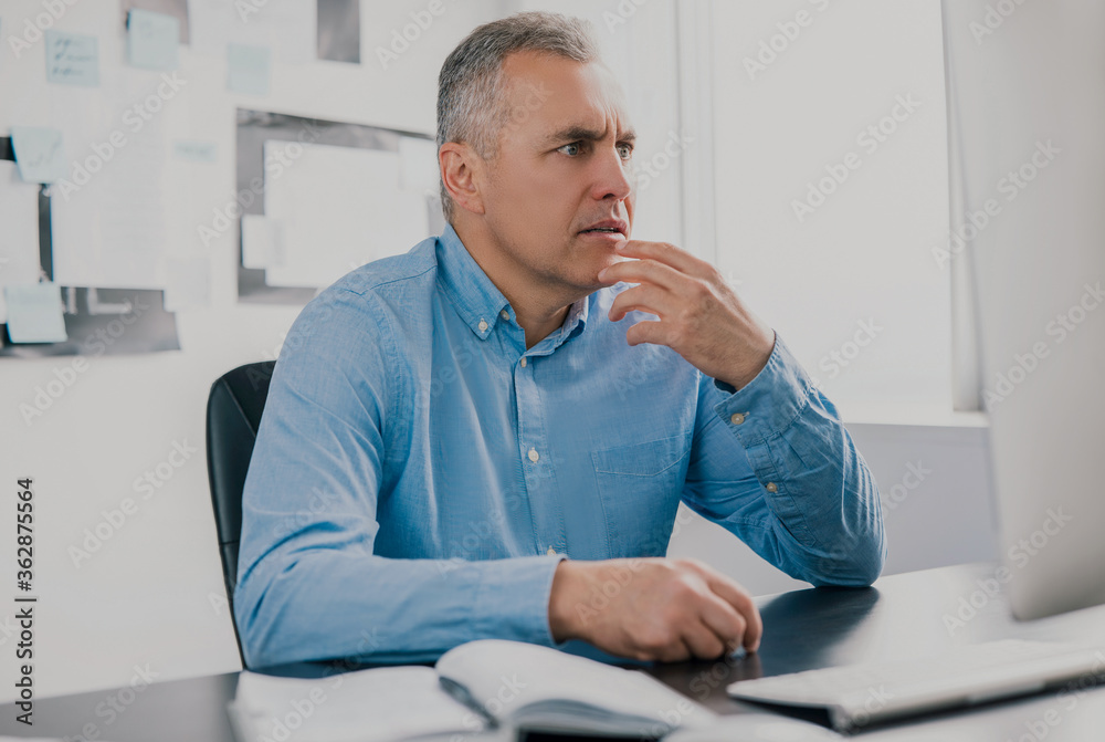 Fototapeta premium handsome gray-haired man sits in his office holding hand near face looking surprised while working on business project, work routine concept