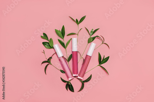 Three lip glosses in different shades of pink decorated with fresh green leaves on pastel pink background. Flat lay style. Organic cosmetics concept. Mockup for your design. Copy space. photo