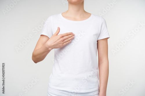 Horizontal photo of young female dressed in casual clothes keeping palm on her chest while meaning word mine on deaf language, isolated over white background © timtimphoto