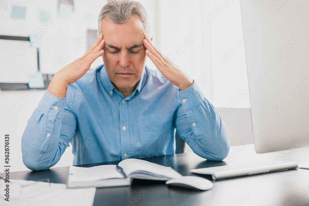 handsome tired gray-haired man sits in his office holding hands near temples looking exhausted while working on business project , stress at workplace concept handsome tired gray-haired man sits in his office holding hands near temples looking exhausted while working on business project , stress at workplace concept