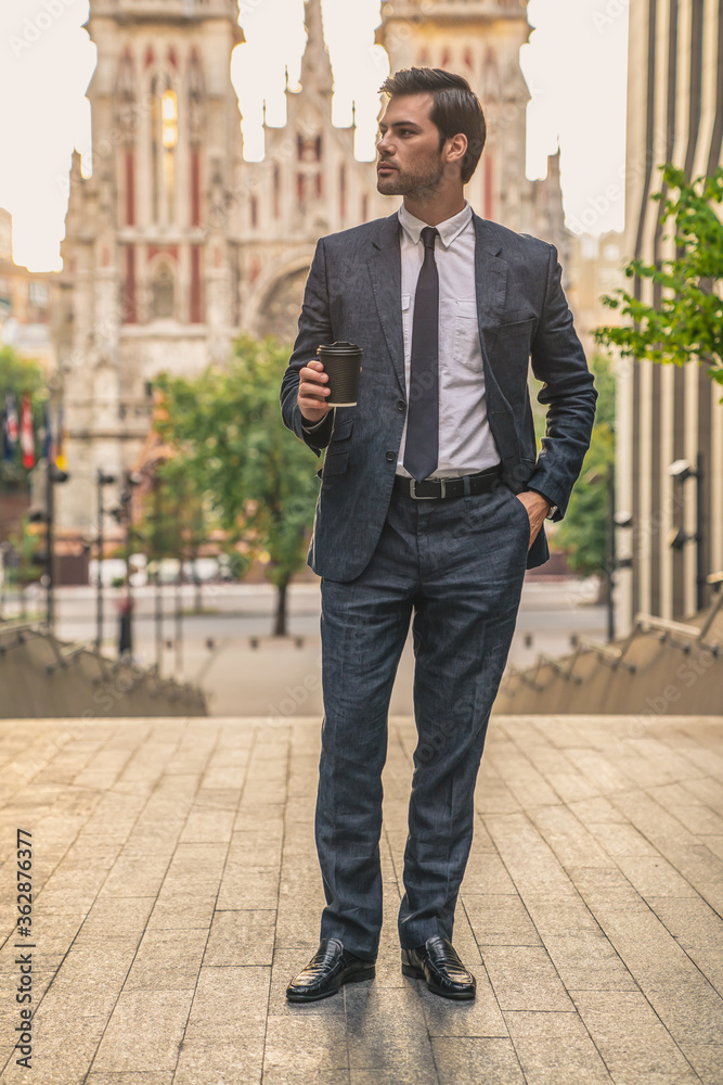 Coffee break. Confident young man in full suit holding coffee cup and looking away while standing outdoors with cityscape in the background Coffee break. Confident young man in full suit holding coffee cup and looking away while standing outdoors with cityscape in the background